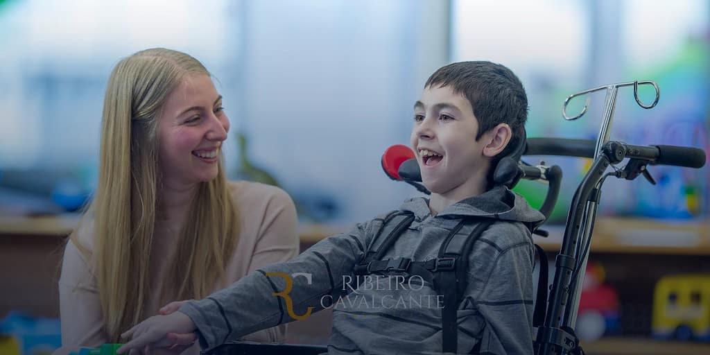 Criança em cadeira de rodas sorrindo ao interagir com mulher loira, ambiente acolhedor e colorido ao fundo.