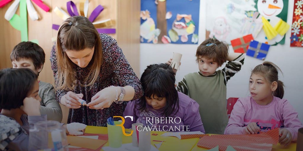 Professora ajudando crianças com atividades artísticas na escola, com desenhos coloridos na parede. Marca: Ribeiro Cavalcante.