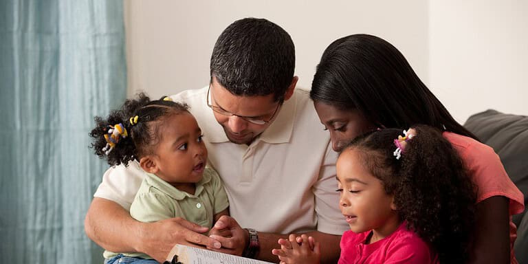 Família unida em momento de oração e leitura, pai, mãe e duas filhas pequenas, em casa, felizes e conectados.