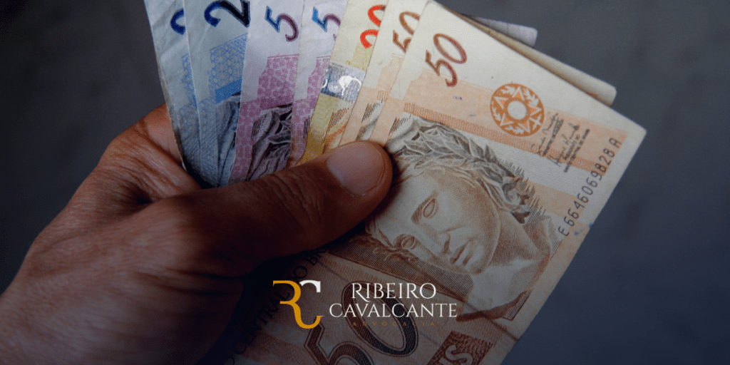Mão segurando notas de real brasileiro, destacando dinheiro e finanças com logo Ribeiro Cavalcante Advocacia.