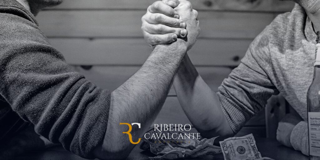 Dois homens participando de uma queda de braço com notas de dinheiro sobre a mesa entre eles. Logo Ribeiro Cavalcante.