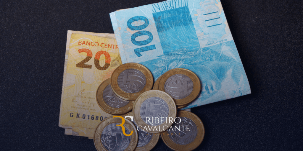 Notas e moedas de real brasileiro em fundo preto com logo da Ribeiro Cavalcante Advocacia.