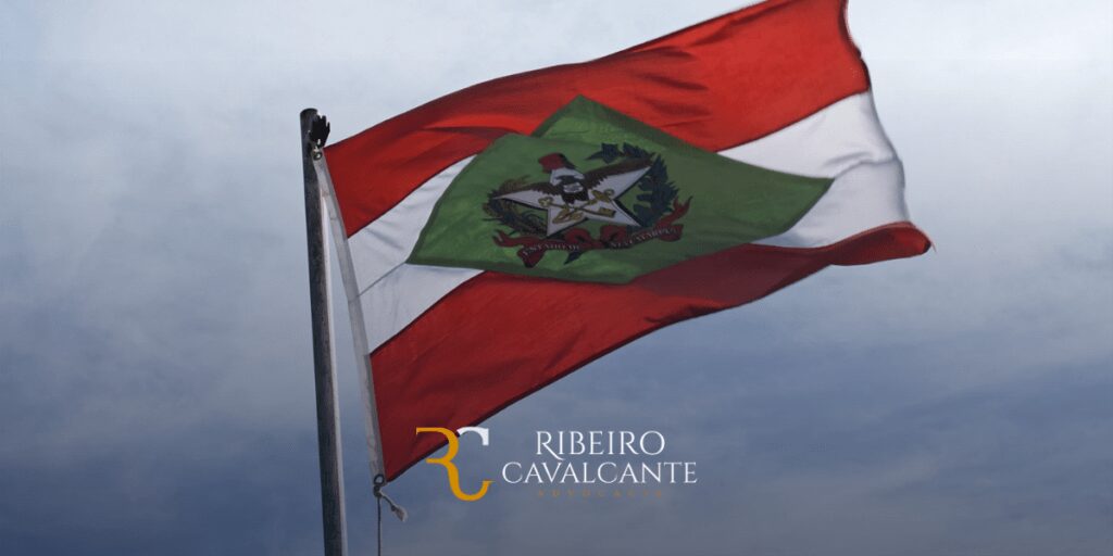 Bandeira do estado de Santa Catarina tremulando ao vento com céu ao fundo, logotipo Ribeiro Cavalcante Advocacia em destaque.