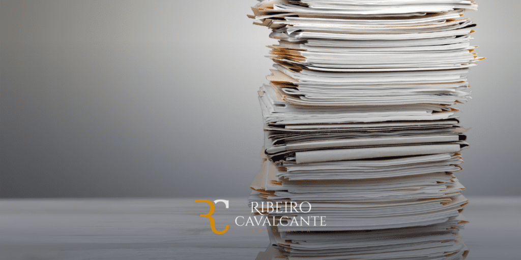 Pilha alta de documentos sobre uma mesa com o logotipo do escritório de advocacia Ribeiro Cavalcante no canto inferior direito.