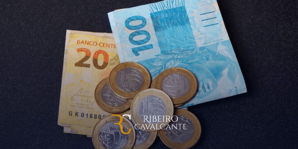 Notas de 20 e 100 reais ao lado de várias moedas de um real em uma superfície escura. Texto: Ribeiro Cavalcante.