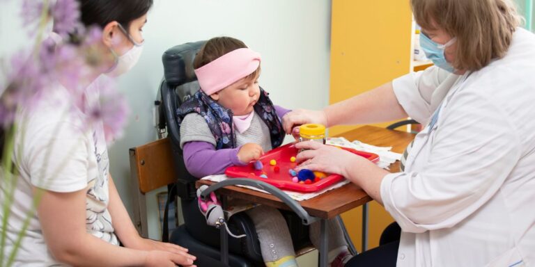 Criança em cadeira de rodas sendo atendida por terapeuta sob supervisão de adulta, representando inclusão e cuidado médico.