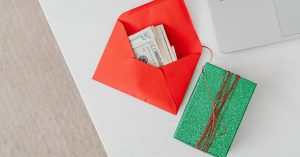 Envelope vermelho com notas de dinheiro ao lado de um presente verde sobre uma mesa de escritório branca. — Foto: www.kaboompics.com