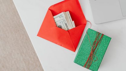 Envelope vermelho com notas de dinheiro ao lado de um presente verde sobre uma mesa de escritório branca. — Foto: www.kaboompics.com