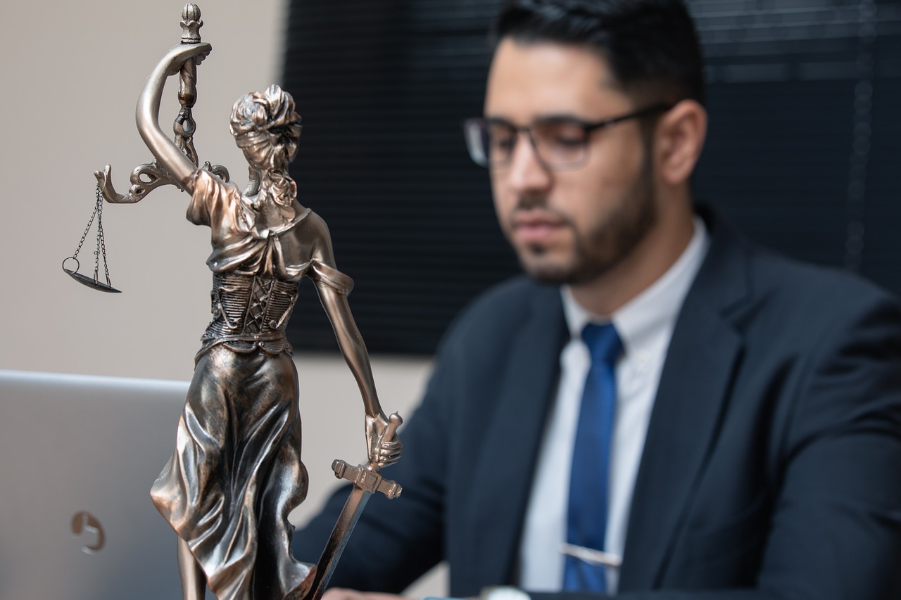 Estátua da deusa Têmis em primeiro plano com advogado desfocado ao fundo trabalhando em um notebook. — Foto: advogadoaguilar