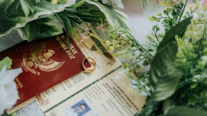 Passaporte vermelho, certidão com foto e aliança de ouro sobre mesa decorada com folhas verdes. — Foto: Rizki Koto