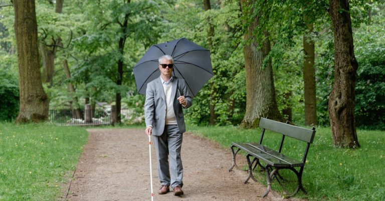 Homem cego caminhando em um parque com bengala guia e guarda-chuva aberto. — Foto: MART PRODUCTION