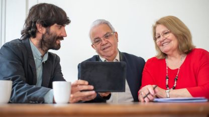 Homem de terno mostra informações em um tablet para um casal de idosos sentados à mesa em um escritório. — Foto: Kampus Production