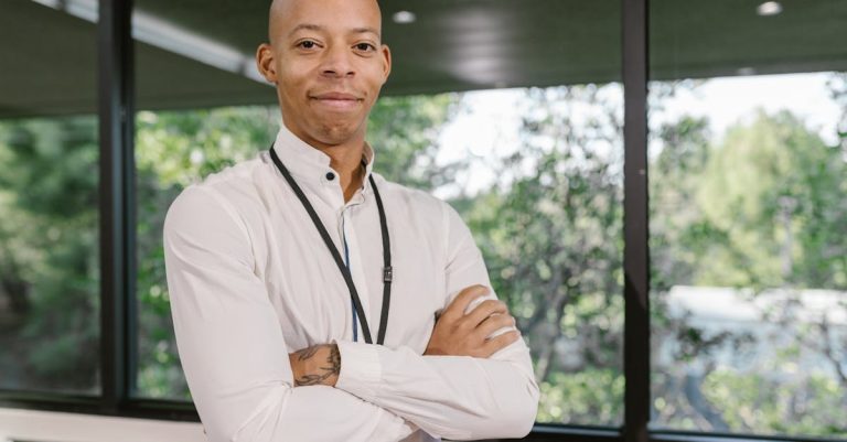 Homem negro de camisa social branca com braços cruzados em um escritório com janelas ao fundo. — Foto: RDNE Stock project
