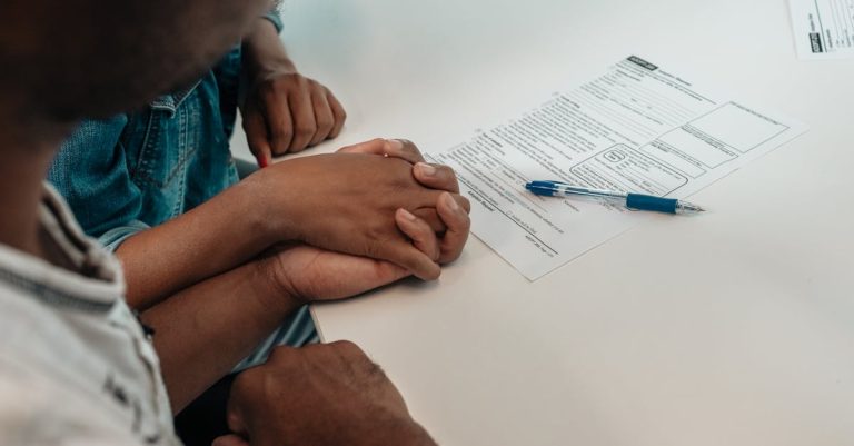 Casal de mãos dadas sobre uma mesa branca com formulários e uma caneta, sugerindo a assinatura de um acordo. — Foto: Kindel Media