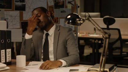 Homem de terno expressando cansaço em um escritório à noite com documentos sobre a mesa. — Foto: cottonbro studio
