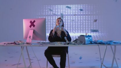 Mulher sentada em mesa com computador, manuseando diversas notas de dinheiro que caem ao seu redor em ambiente moderno. — Foto: Tima Miroshnichenko