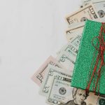 Cédulas de dólar espalhadas sob uma pequena caixa de presente verde com laço vermelho em fundo branco. — Foto: www.kaboompics.com