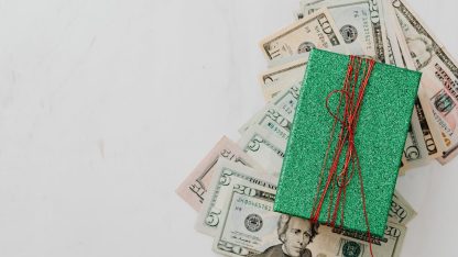 Cédulas de dólar espalhadas sob uma pequena caixa de presente verde com laço vermelho em fundo branco. — Foto: www.kaboompics.com