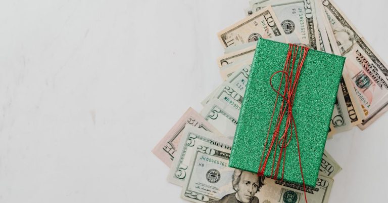Cédulas de dólar espalhadas sob uma pequena caixa de presente verde com laço vermelho em fundo branco. — Foto: www.kaboompics.com