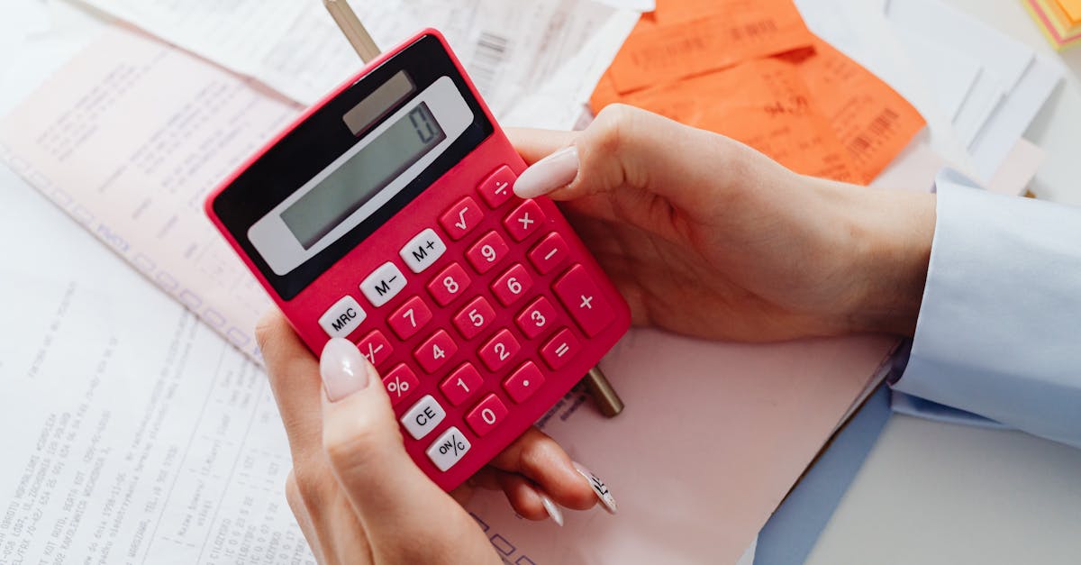 Mãos de uma pessoa segurando uma calculadora rosa sobre documentos e boletos em uma mesa de escritório. — Foto: www.kaboompics.com