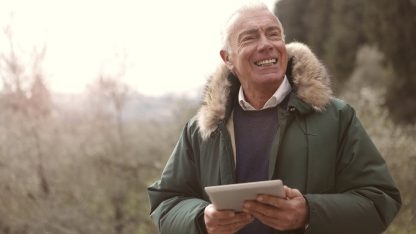 Homem idoso sorridente usando um tablet ao ar livre vestindo casaco de inverno. — Foto: Andrea Piacquadio