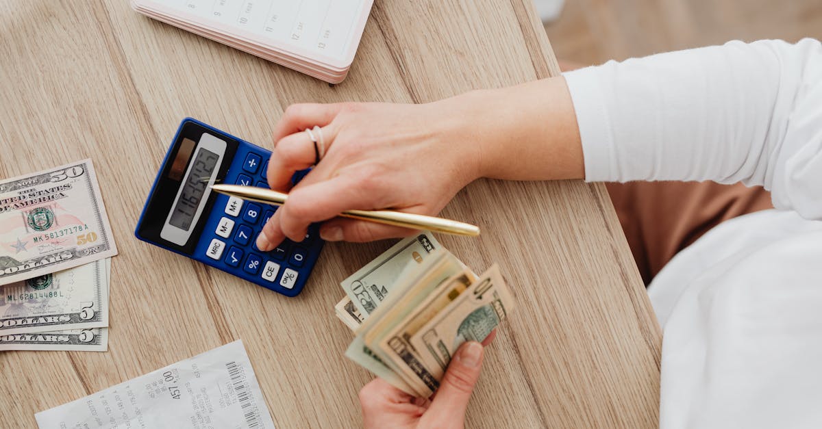 Pessoa usando calculadora e segurando notas de dinheiro sobre uma mesa de madeira. — Foto: www.kaboompics.com