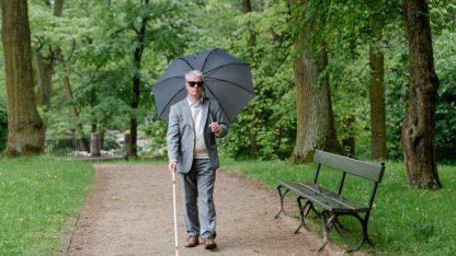 Homem cego caminhando em um parque com bengala guia e guarda-chuva aberto. — Foto: MART  PRODUCTION
