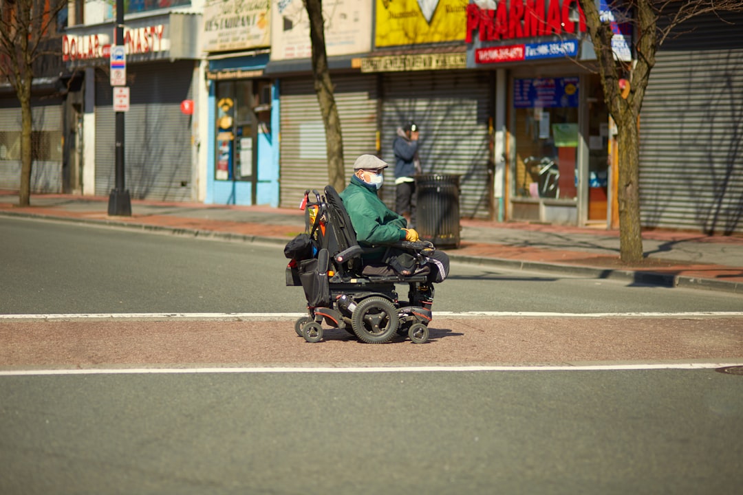 Homem idoso em cadeira de rodas motorizada atravessando uma rua urbana em frente a comércios. — Foto: redens desrosiers