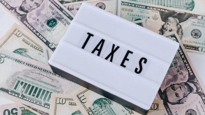 Placa branca com a palavra TAXES escrita em preto sobre diversas notas de dólar americano. — Foto: www.kaboompics.com