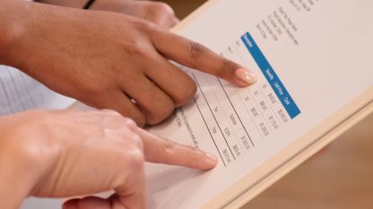 Close de duas pessoas apontando para valores em uma fatura ou documento financeiro sobre uma prancheta. — Foto: Kindel Media