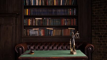 Estátua da justiça e martelo de juiz sobre mesa verde em escritório de advocacia com estante de livros ao fundo. — Foto: KATRIN  BOLOVTSOVA