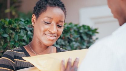 Mulher sorridente recebendo ou entregando um envelope pardo e uma caixa de papelão para outra pessoa. — Foto: Kindel Media