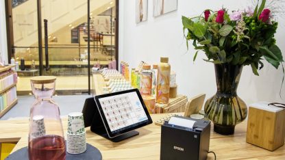 Balcão de loja com tablet de vendas, impressora de recibos, garrafas e um vaso de flores ao fundo. — Foto: Tom Tillhub
