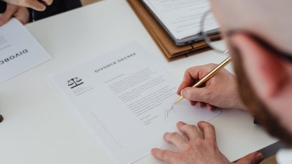 Pessoa assinando documento jurídico com símbolo da balança da justiça sobre uma mesa branca. — Foto: www.kaboompics.com