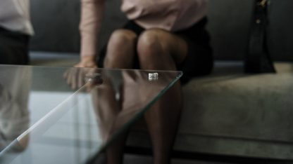 Par de alianças de casamento sobre uma mesa de vidro com uma mulher sentada ao fundo em ambiente de escritório. — Foto: cottonbro studio