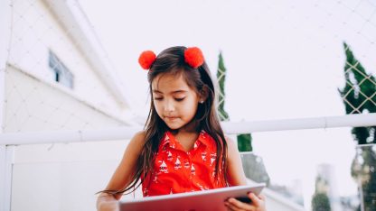 Menina de blusa vermelha segurando e olhando para um tablet em uma área externa com rede de proteção. — Foto: Helena Lopes