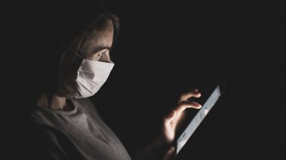 Pessoa usando máscara de proteção facial manuseando um tablet no escuro, com a tela iluminando seu rosto. — Foto: Engin Akyurt