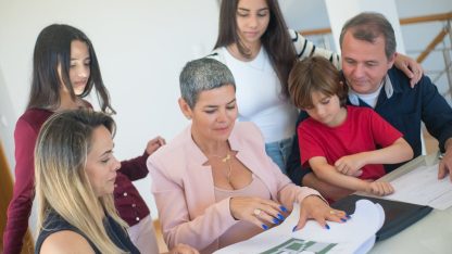 Família reunida em volta de uma mesa analisando documentos e plantas com uma profissional em um ambiente iluminado. — Foto: Kampus Production