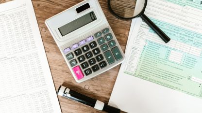 Calculadora branca, lupa e formulários de impostos sobre uma mesa de madeira. — Foto: RDNE Stock project