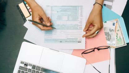 Vista superior de mãos preenchendo formulário de declaração financeira sobre mesa com notebook, óculos e dinheiro. — Foto: Nataliya Vaitkevich