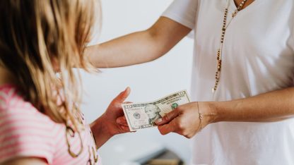 Uma mulher entregando uma nota de dólar para uma criança pequena em um ambiente doméstico. — Foto: www.kaboompics.com
