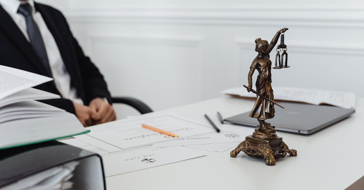 Estátua da justiça sobre mesa de escritório com advogado ao fundo e documentos jurídicos espalhados. — Foto: www.kaboompics.com