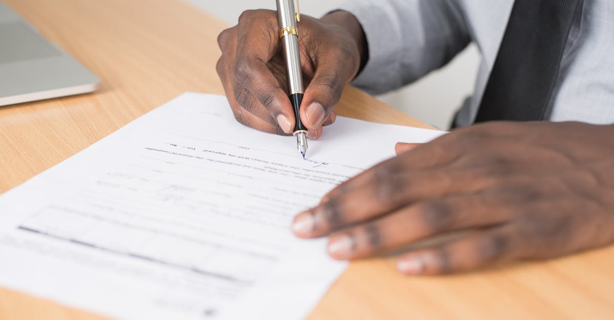Close das mãos de um homem negro assinando um documento oficial com uma caneta tinteiro sobre uma mesa de madeira. — Foto: Cytonn Photography