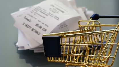 Miniatura de carrinho de compras dourado em frente a uma pilha de notas fiscais e recibos de papel. — Foto: Alexas_Fotos