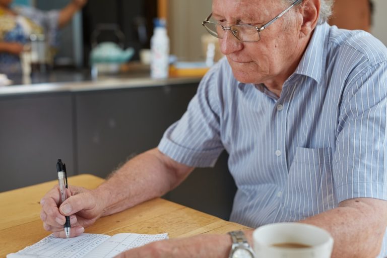 Homem idoso de óculos escrevendo em um caderno sobre uma mesa de madeira com uma xícara de café ao lado. — Foto: Sweet Life