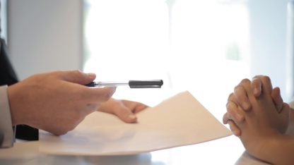 Pessoa em traje social entregando uma caneta e papel para outra pessoa assinar sobre uma mesa branca. — Foto: Andrea Piacquadio