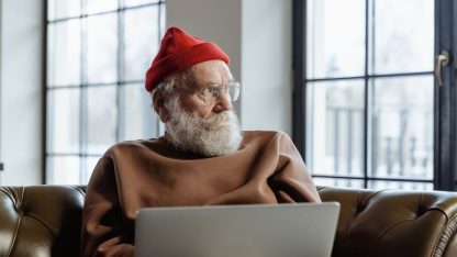 Homem idoso de barba branca e touca vermelha usando notebook sentado em um sofá de couro. — Foto: MART  PRODUCTION
