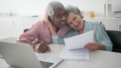 Casal de idosos sorridentes analisando papéis e documentos em frente a um notebook em um escritório iluminado. — Foto: Kampus Production