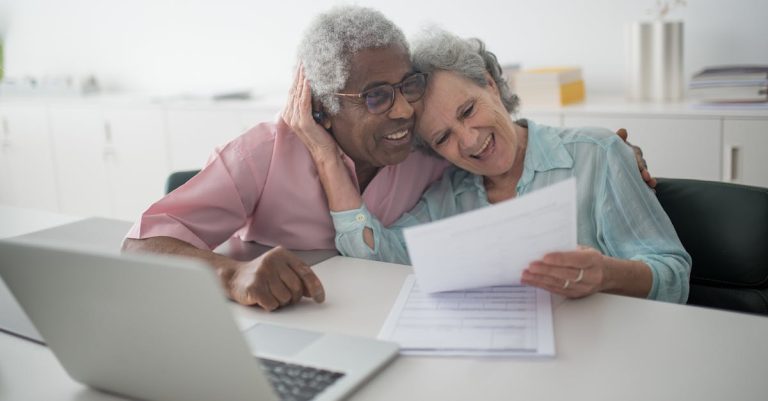 Casal de idosos sorridentes analisando papéis e documentos em frente a um notebook em um escritório iluminado. — Foto: Kampus Production