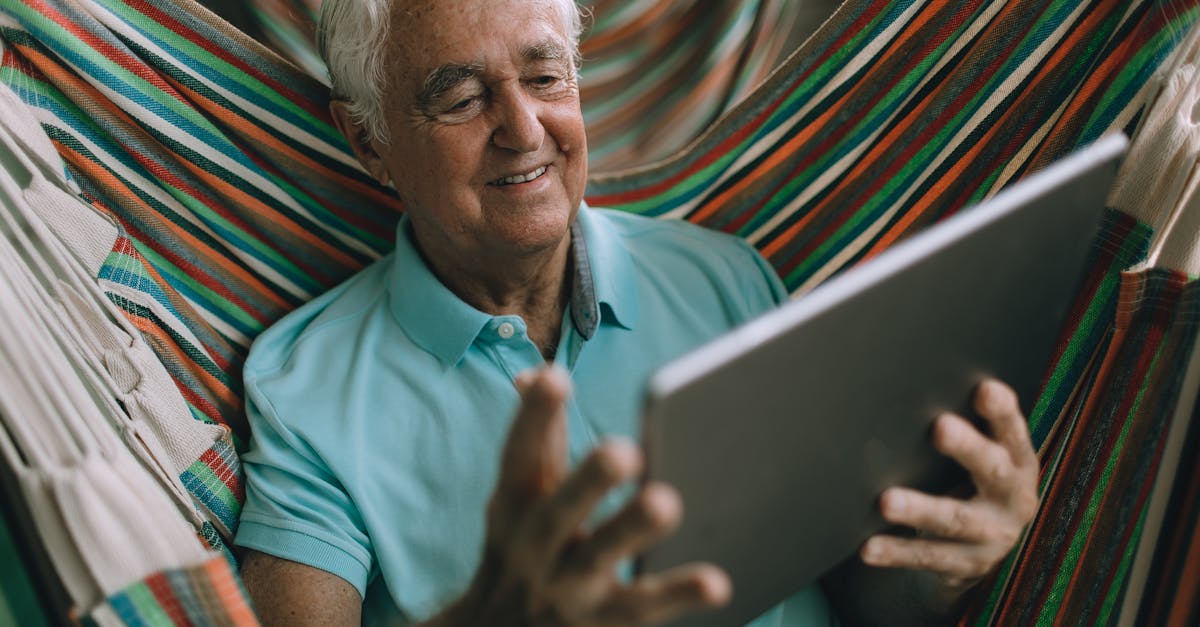 Homem idoso sorridente sentado em uma rede colorida enquanto utiliza um tablet digital. — Foto: Helena Lopes